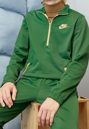 Osoba ubrana w zielony dres Nike z beżowymi detalami na zamkach siedzi na drewnianej skrzyni na tle jasnoniebieskiej poduszki.
