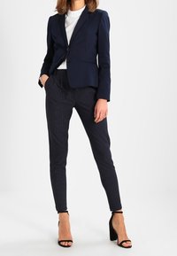 Granatowa, dopasowana marynarka na jeden guzik, zestawiona z ciemnymi spodniami w prążki. Całość dopełniają czarne sandały na obcasie z paskami.