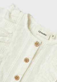 Top de algodón blanco con cierre de botones, detalles texturizados y acentos con volantes en el escote y los hombros. Incluye botones de madera.