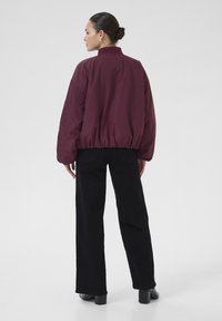 Veste bomber bordeaux avec un col côtelé, une taille élastique et des manches longues, assortie à un pantalon noir à jambe large et des talons.