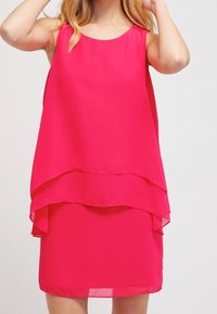 Kobieta ubrana w bezrękawową, jasnoróżową, warstwową sukienkę z okrągłym dekoltem, stojąca na jednolitym jasnym tle.