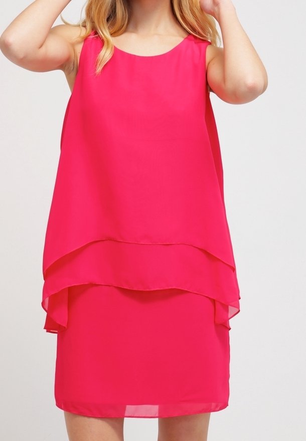 Kobieta ubrana w bezrękawową, jasnoróżową, warstwową sukienkę z okrągłym dekoltem, stojąca na jednolitym jasnym tle.