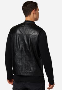 Schwarze Lederweste mit einer glatten Textur, taillierter Passform und ohne Verschlüsse. Hat eine Rückenpasse und Nähte an den Panels. Getragen über einem schwarzen Langarmhemd.