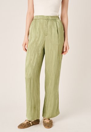 Mujer con pantalones de pierna ancha de cintura alta, color verde claro con textura, con bolsillos laterales y zapatos planos dorados, combinados con una camiseta sin mangas blanca de canalé.