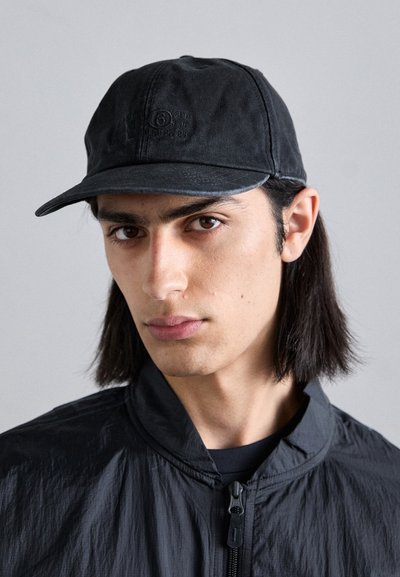 Casquette noire en tissu texturé avec une visière incurvée ; présente des détails de logo en relief à l'avant. Portée avec une veste noire zippée.
