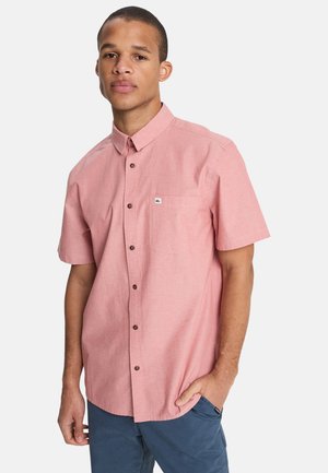 Koszula na krótki rękaw na guziki z jasnoróżowego materiału, z kieszenią na piersi i brązowymi guzikami. Małe logo na kieszeni. Luźny krój.