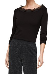 Kvinna iklädd en svart långärmad tröja med volangkrage och högmidjade svarta byxor med silverprickigt mönster.