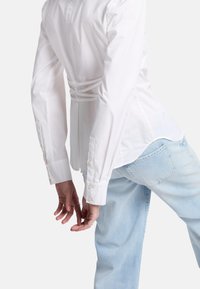 Vit bomullsskjorta med en tight baksida och knytdetaljer, långa ärmar, knäppta manschetter och en rundad fåll. Matchas med ljusblå jeans.