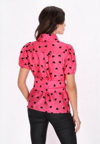 Lyserød skjorte med sort hjerte mønster, korte puffærmer og belted talje. Stoffet har en let skinnende tekstur, og kraven er fremtrædende.