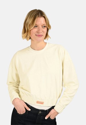 Femme aux cheveux blonds courts portant un sweat-shirt court crème à manches longues et un jean noir, les mains dans les poches, regardant vers la droite.