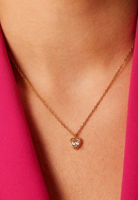 Gouden ketting met een hartvormige hanger met een transparante steen, gedragen tegen een huidskleurige achtergrond met een roze stof.
