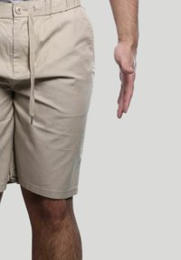 Pantaloni beige con una vita elastica, chiusura con cordoncino e un tessuto leggero e informale. Cucitura laterale visibile e gesto della mano al lato.