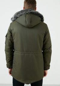 Parka verde olivo con acabado de piel sintética en el capucho. Cuenta con un cierre de cremallera, bolsillos laterales y un dobladillo curvado en la parte posterior. Textura suave, diseño aislante.