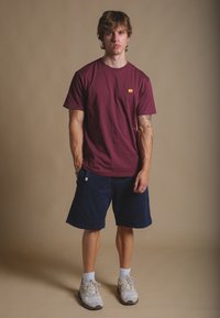 Maglietta di cotone bordeaux con logo di un elefante arancione, abbinata a pantaloni corti blu navy e scarpe da ginnastica grigie con dettagli bianchi e calzini a metà polpaccio.