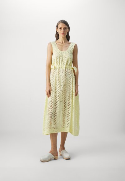 Robe en dentelle jaune avec un décolleté carré et une ceinture à la taille, dotée d'un superposé semi-transparent, complétée par des sabots blancs à enfiler.