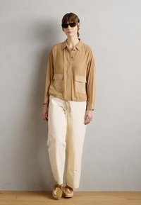 Light brown shirt jacket with two front pockets, paired with cream-colored trousers. Soft fabric and relaxed fit, complementing tan shoes.