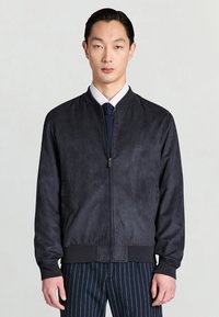 Giacca bomber nera con texture liscia simile a camoscio, polsini e orlo a coste, e cerniera frontale. Indossata con camicia bianca e cravatta a fantasia.
