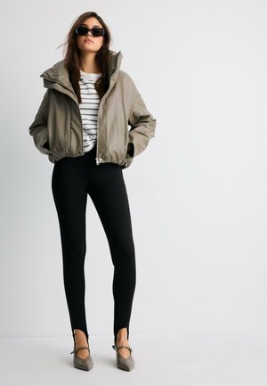 Femme portant une veste beige oversize, une chemise rayée, des leggings noirs, des chaussures pointues et des lunettes de soleil noires, debout devant un fond blanc.