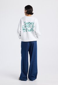 Persona con cabello corto y oscuro, vistiendo un sudadera blanca con el texto "Sunday Coffee Club" y gráficos de tazas, combinada con unos pantalones vaqueros oscuros y holgados.