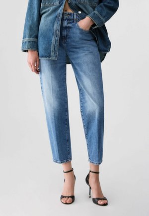 Donna con jeans a vita alta azzurri chiari e giacca di jeans, in piedi con una mano in tasca e tacchi alti neri con cinturino alla caviglia.