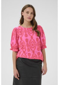 Blusa rosa con maniche a palloncino e ricami floreali arancioni. Indossata su una gonna di pelle nera. Tessuto liscio, vestibilità rilassata.