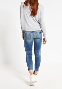 Suéter gris claro combinado con jeans skinny azules que presentan áreas desgastadas y bolsillos traseros, luciendo con zapatillas blancas de caña alta.