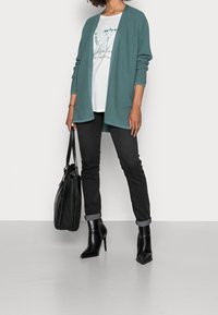 Mujer vestida con un cárdigan de canalé en color teal, camiseta gráfica blanca, pantalones negros remangados, botas negras de tacón, sosteniendo un bolso de cuero negro a un lado.