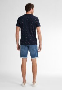 Laiva sininen lyhythihainen paita, jossa on punaisia kuvioita, yhdistettynä vaaleisiin farkkushortsit. Paidassa on pyöreä kaula-aukko ja shortseissa on käännetty helma.