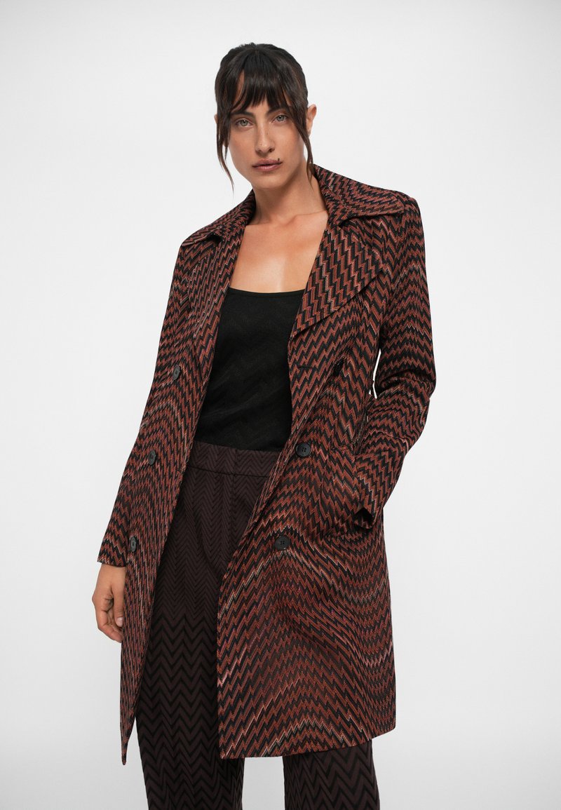 Manteau à motif zigzag marron et noir avec un col, boutons double boutonnage et manches longues, assorti d'un haut et d'un pantalon noirs.