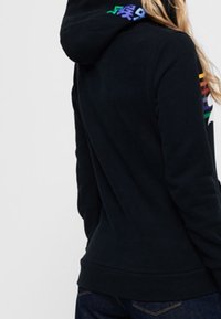 Persona con un cappuccio nero che presenta lettere colorate sul cappuccio e strisce arcobaleno sul petto, mostrata di spalle e di lato.