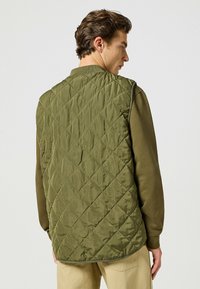 Gilet imbottito verde oliva con motivo a diamante, caratterizzato da un colletto arrotondato e design senza maniche, indossato sopra una camicia a maniche lunghe.