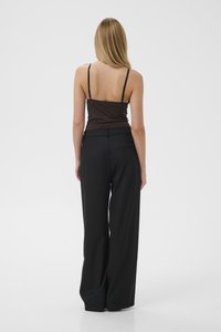 Donna con lunghi capelli biondi che sta in piedi di spalle, indossando un top senza maniche di colore marrone scuro e pantaloni larghi neri su uno sfondo bianco.