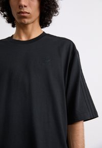 Sort bomulds-t-shirt med korte ærmer, rund hals og præget grønt Adidas-logo på brystet med tre-stribe detalje på skuldrene.