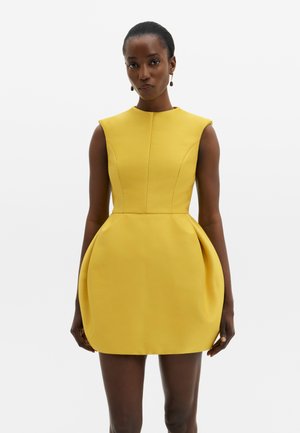 Femme portant une robe jaune sans manches, structurée avec des hanches rembourrées, debout devant un fond blanc uni.