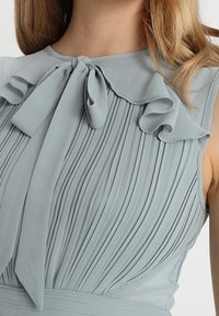 Vaaleansininen mekko, jossa on poimutettu kangasmalli, röyhelökaulus ja solmittu nauhadetalji kaula-aukossa. Sujuva pinta.