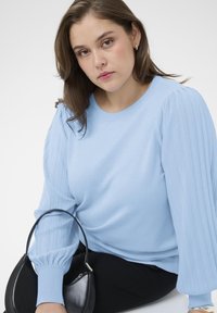 Világoskék kötött pulóver bordázott ballon ujjakkal, fekete kézitáskával párosítva, amelynek ívelt kialakítása és ezüst cipzára van.