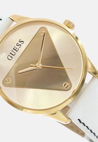 Gouden analoge watch met een driehoekige wijzerplaat, witte leren band en gouden accenten. Beschikt over drie wijzers en de merknaam "GUESS."