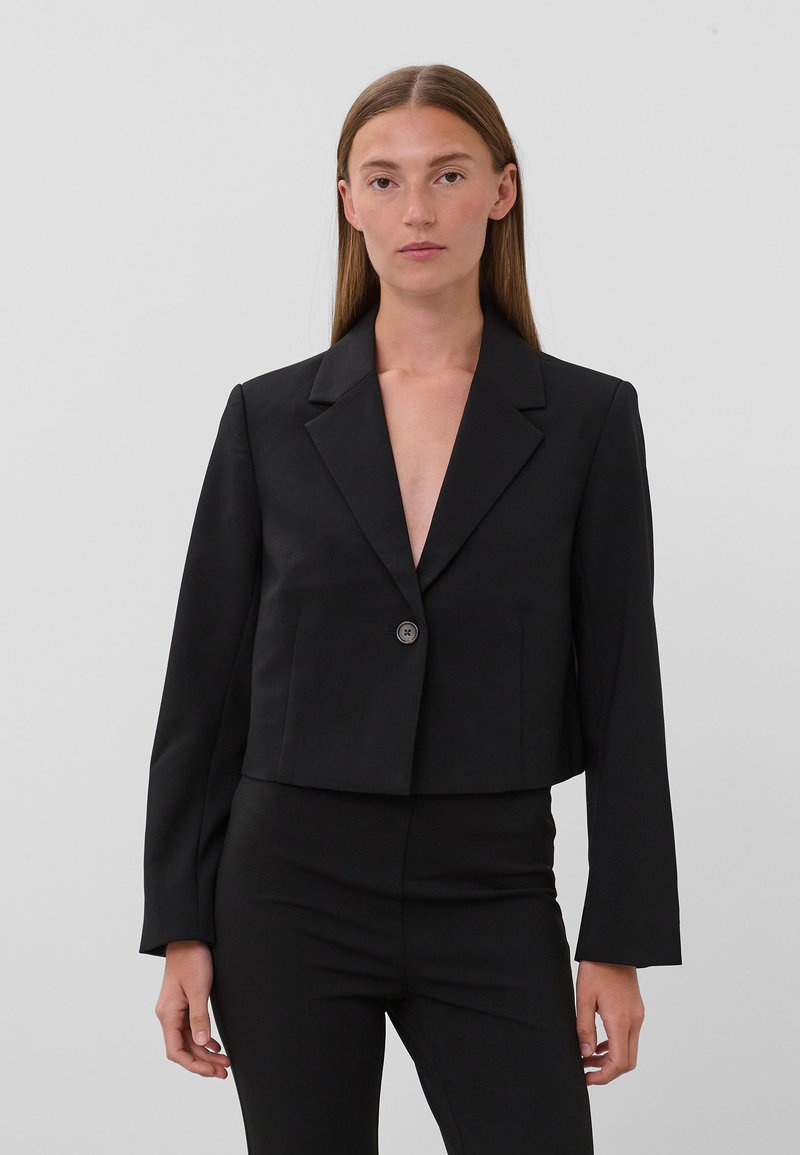 Vrouw met steil bruin haar, die een nauwsluitend zwart colbert met één knoop en bijpassende zwarte broek draagt tegen een effen grijze achtergrond.