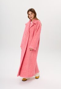 Rosa übergroße Jacke aus strukturiertem Stoff, mit Reverskragen, zwei großen Frontknöpfen und weit geschnittenen Ärmeln. Tragen Sie sie über cremefarbenen Hosen und gelben Schuhen.