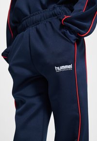 Marinträningsbyxor med röd sidopiping, elastisk midja, slim fit och en vit "hummel PERFORMANCE"-logotyp nära fickan.