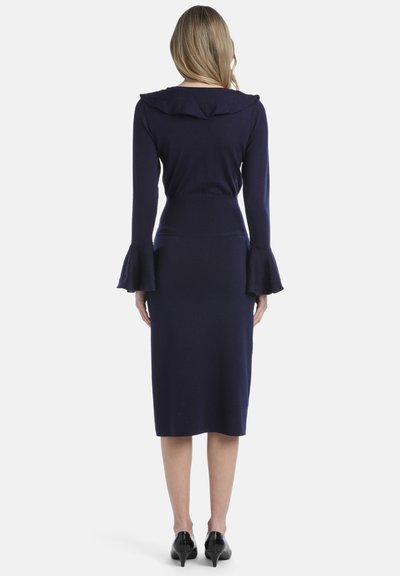 Mornarsko pleteno oblačilo s kapuco, prilegajoča silhueta, dolgi rokavi, razširjeni manšeti in dolžina do kolen, v kombinaciji s črnimi petkami.