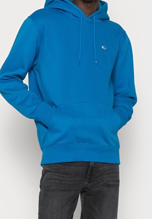 Mężczyzna ubrany w jaskrawoniebieską bluzę z kapturem, z kieszenią z przodu i małym logo na piersi, w połączeniu z czarnymi dżinsami.