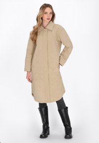 Beige quiltet jakke med krave, sidelommer og lige silhuet, parret med sorte støvler til knæet. Glat tekstur og varmt design.