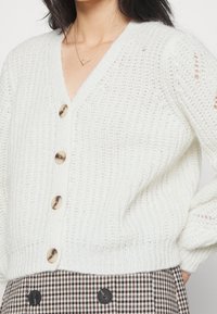 Vit stickad kofta med V-ringning, prydd med stora beige knappar och öppna stickdetaljer. Texturerad tyg med en kortad och avslappnad passform.