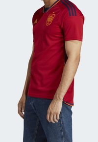 Červený fotbalový dres s modrými a žlutými akcenty, s krátkými rukávy, kulatým výstřihem a logem týmu na hrudi. Vyrobený z prodyšné látky.