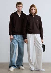 Καφέ φούτερ με φερμουάρ και μικρό λογότυπο, συνδυασμένο με άνετο ανοιχτό μπλε τζιν και λευκά παπούτσια. Το μοντέλο κρατά μια μικρή μαύρη τσάντα.