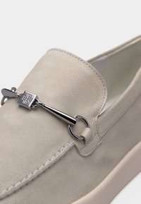Μπεζ μοκασίνι από σουέντ με ασημένια μεταλλική λεπτομέρεια τύπου bit που διαθέτει τετράγωνο έμβλημα και στρογγυλές διακοσμήσεις πάνω στο επάνω μέρος.