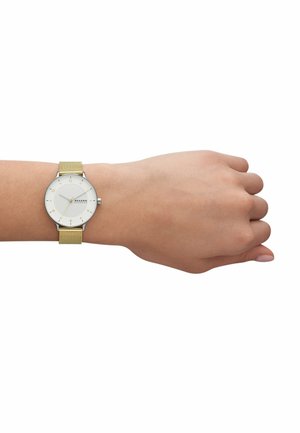 Orologio argento con cassa rotonda, quadrante bianco, indicatori delle ore dorati e cinturino a rete dorato. Indossato su una mano umana, che mette in mostra il suo design.