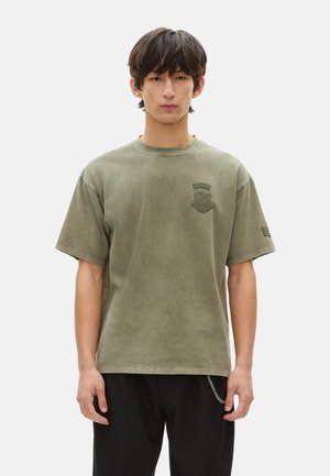 Olivegrünes, oversized Baumwoll-T-Shirt mit runder Halslinie, kurzen Ärmeln und einem gestickten Emblem auf der Brust. Weiche, glatte Textur.