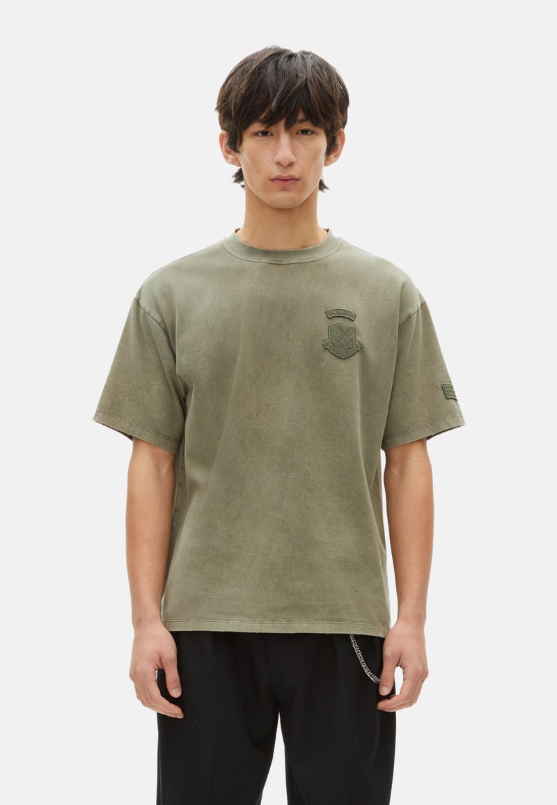 Camiseta de algodón de color verde oliva, de corte oversized, con cuello redondo, mangas cortas y un emblema bordado en el pecho. Textura suave y lisa.